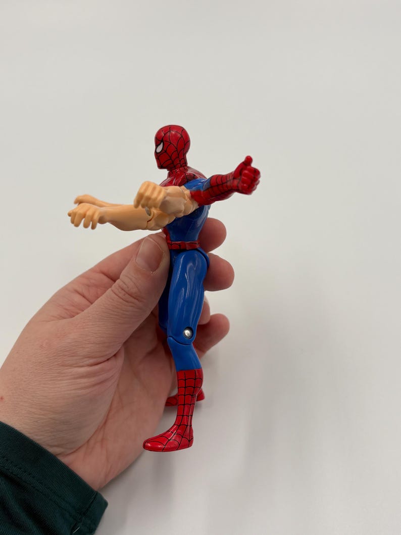 May include: A Spider-Man action figure in a dynamic pose. The figure features a red and blue costume with a red mask. The figure's arms are extended, with one hand in a thumbs-up gesture. The figure is held against a white background.