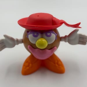 May include: A plastic toy potato head with a red hat and orange shoes. The toy is made of red, white, and purple plastic and has a purple mouth and yellow nose.