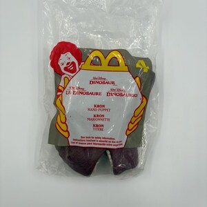 May include: A sealed plastic bag containing a Kron hand puppet from the Disney movie Dinosaur. The packaging features the McDonald's logo, the movie title in multiple languages, and the puppet's name. The puppet is a dark maroon color.