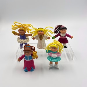 Peut inclure: Cinq figurines vintage Cabbage Patch Kids. Les poupées portent diverses tenues, dont une ballerine, un ensemble d'hiver et une robe. Les poupées ont des cheveux en fil jaune, marron et brun foncé. Les poupées sont sur un présentoir en acrylique transparent.