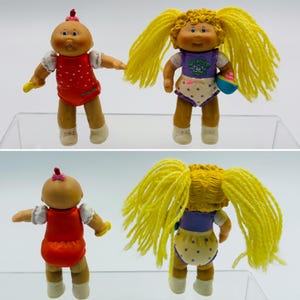 May include: Two small dolls with tan skin and white shoes. One doll has a bald head with a small pink bow, wearing a red polka-dot outfit. The other doll has long yellow yarn hair, wearing a purple top and white polka-dot shorts with a blue bowl.