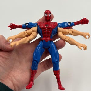 May include: A Spider-Man action figure with multiple arms, in a Vitruvian Man pose. The figure is primarily red and blue, with a black spider emblem on the chest. The figure's arms are tan and blue.