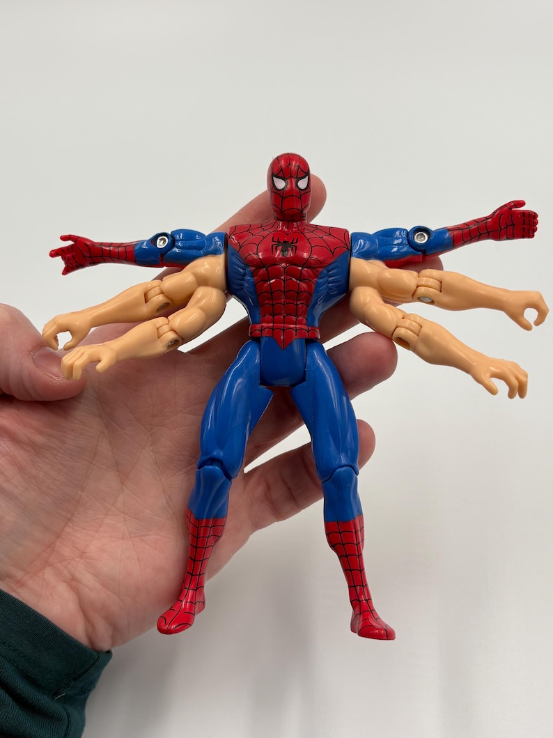 May include: A Spider-Man action figure with multiple arms, in a Vitruvian Man pose. The figure is primarily red and blue, with a black spider emblem on the chest. The figure's arms are tan and blue.