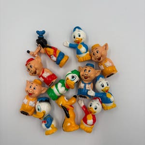 May include: A collection of 11 small plastic figurines of Disney characters, including Mickey Mouse, Donald Duck, Goofy, and the three little pigs. The figurines are brightly colored and feature various poses.