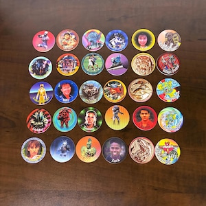 May include: A collection of thirty circular Power Rangers pogs arranged in five rows of six. Each disc features colorful illustrations of characters, including Yellow Ranger, Billy, Tommy, Zack, Trini, Jason, Goldar, Finster, and various Zords.