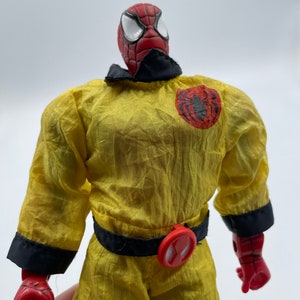 Puede incluir: Una figura de acci&oacute;n roja y negra de Spider-Man con un mono amarillo, cintur&oacute;n negro y hebilla roja. La figura tiene un logotipo de ara&ntilde;a negro en el pecho y un logotipo de ara&ntilde;a rojo en la hebilla del cintur&oacute;n.