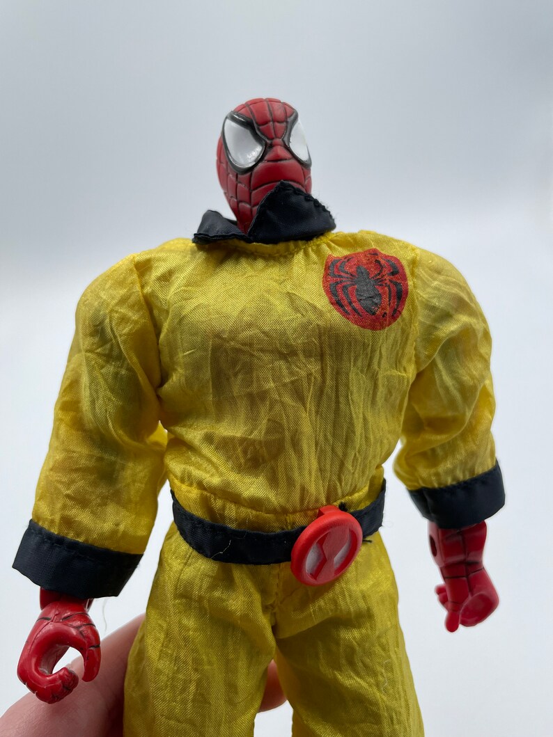 Puede incluir: Una figura de acci&oacute;n roja y negra de Spider-Man con un mono amarillo, cintur&oacute;n negro y hebilla roja. La figura tiene un logotipo de ara&ntilde;a negro en el pecho y un logotipo de ara&ntilde;a rojo en la hebilla del cintur&oacute;n.