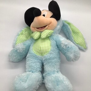 K&ouml;nnte beinhalten: Ein blauer Pl&uuml;sch-Mickey Mouse, der ein gr&uuml;nes und wei&szlig;es Gingham-Kaninchenkost&uuml;m tr&auml;gt. Die Hasenohren sind lang und schlaff.