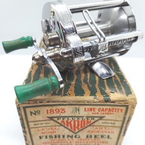 May include: A vintage Pflueger Akron fishing reel, model 1893, with a silver finish and green handles. The reel sits atop its original, aged cardboard box with text including "Akron" and "Fishing Reel".