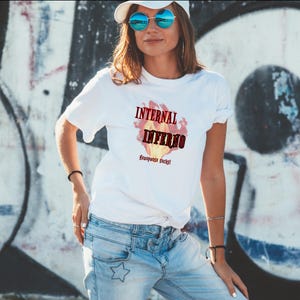 Könnte beinhalten: Weißes T-Shirt mit dem Text "INTERNAL INFERNO" über einer Flammen-Grafik. Darunter steht "Menopause sucks!". Das Shirt wird mit hellblauen Jeans und einer weißen Kappe getragen.