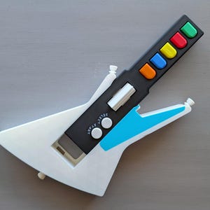 Puede incluir: Una guitarra de juguete de plástico blanco con un cuerpo de plástico negro y azul y un mástil de plástico negro con una fila de botones de colores. Los botones son rojo, naranja, amarillo, verde, azul y morado. La guitarra tiene un interruptor de plástico blanco con la etiqueta "SELECT" y "START" y una perilla de plástico blanco con la etiqueta "1/2/3/C".