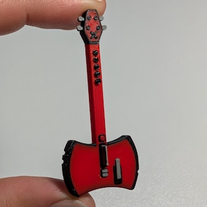 May include: A small, red and black guitar-shaped object with an axe-like body design. The headstock features four tuning pegs, and the long neck includes five circular fret markers. The body has a central bridge and a rectangular pickup detail.