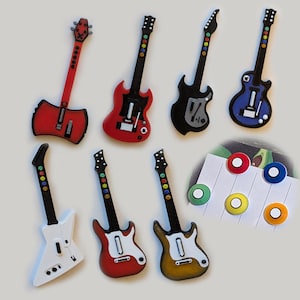 May include: A collection of seven miniature guitar-shaped game controllers in various colors including red, black, blue, white, and gold, alongside five circular buttons in red, blue, green, yellow, and orange arranged on a white paper strip.
