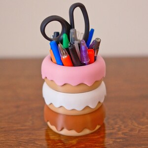 Puede incluir: Un organizador de escritorio de varios niveles con forma de donut hecho de pl&aacute;stico rosa, marr&oacute;n y blanco. El organizador est&aacute; lleno de varios bol&iacute;grafos, l&aacute;pices y un par de tijeras.