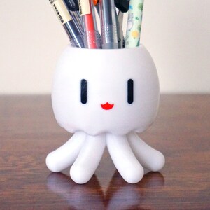 May include: White octopus-shaped desk organizer with a smiling face, holding a variety of pens and pencils, including a green pen with the text "Nightbloom" on it.