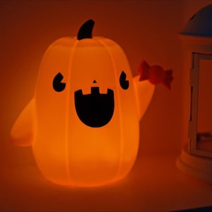 May include: A glowing orange pumpkin with a friendly face and a candy cane in its hand. The pumpkin is lit from within and casts a warm glow. A white lantern with a glass window is in the background.