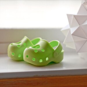 May include: Two bright green plastic clogs, one slightly larger than the other, are sitting on a white surface.