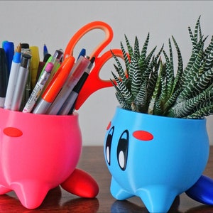 May include: Two 3D printed character planters, one pink and one blue, with a cartoon face. The pink planter is filled with pens and pencils, while the blue planter holds a green succulent plant.