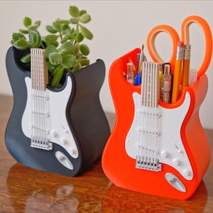 May include: Two guitar-shaped desk tidies, one black and one orange. The black guitar holds a small plant, while the orange guitar holds pencils, scissors, and other stationery. Both have white pickguards and silver hardware.