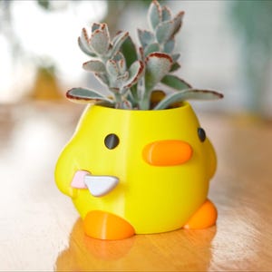 Puede incluir: Una maceta amarilla con forma de pato con ojos negros, pico naranja y patas. La maceta contiene una planta suculenta con hojas verdes y rojizas. Un pequeño cuchillo blanco y rosa está unido al costado del pato. La maceta está sobre una superficie de madera.