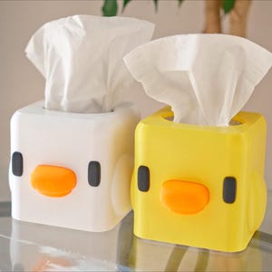Può includere: Due copri scatola di fazzoletti a forma di pulcini dei cartoni animati. Uno è bianco con un becco arancione e occhi neri. L'altro è giallo con un becco arancione e occhi neri.