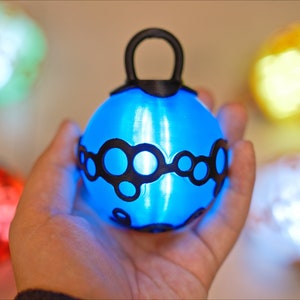 May include: A glowing blue sphere with a black loop handle and a black circular design is held in a hand. Other glowing spheres in green, red, yellow, and white are in the background.