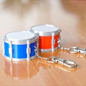 Può includere: Due portachiavi a forma di batteria in miniatura. Uno è blu, l'altro rosso, entrambi con dettagli argentati e pelli bianche. Ogni portachiavi ha una chiusura argentata. I portachiavi sono su una superficie di legno.