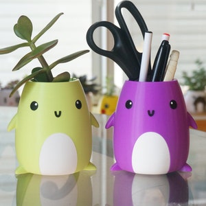 May include: Two cute penguin-shaped planters, one yellow with a green succulent plant and the other purple with a white and black pen holder.