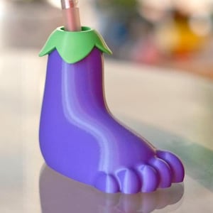 May include: Purple eggplant-shaped pen holder with a green top and a pen inside. The holder is shaped like a foot with five toes.