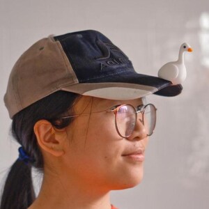 May include: A baseball cap with a tan and navy blue design, featuring a small white goose figurine perched on the brim. The cap is worn by a person wearing glasses and an orange shirt.