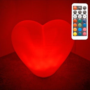 May include: A large, white heart-shaped LED light with a remote control. The light is currently glowing red. The remote control has buttons for different colours, including red, orange, yellow, green, blue, indigo, and violet.