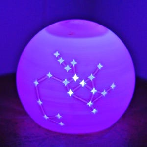 May include: A glowing, spherical lamp with a constellation design. The lamp emits a soft purple light, with a constellation pattern of white stars and connecting lines. The lamp is placed in a dark corner.