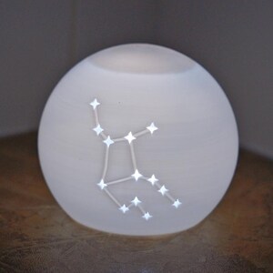 May include: A glowing white orb-shaped lamp with a constellation design. The constellation is illuminated with small, bright lights. The lamp sits on a wooden surface, and the background is a neutral color.
