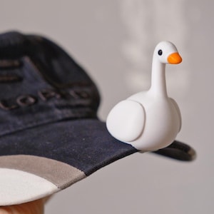 May include: A baseball cap with a white brim, gray and navy blue panels, and the word "FLORIDA" embroidered on the front. A small white goose figurine with an orange beak is perched on the cap's brim.