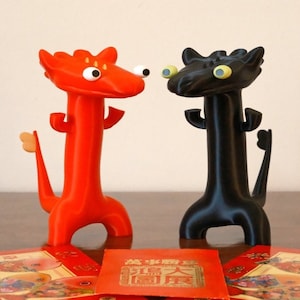 May include: Two small, 3D printed dragon figurines, one red and one black, stand facing each other on a wooden table.  Red envelopes with Chinese characters are in front of the dragons.