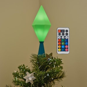 May include: A small artificial Christmas tree with star ornaments and a green, diamond-shaped tree topper. A remote control with colour options is included. The tree is decorated with silver tinsel and star-shaped ornaments.