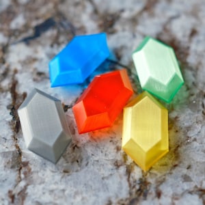 Peut inclure: Cinq gemmes en plastique de forme octogonale de différentes couleurs : gris, bleu, rouge, jaune et vert. Les gemmes sont disposées en grappe sur une surface texturée de couleur brun clair.