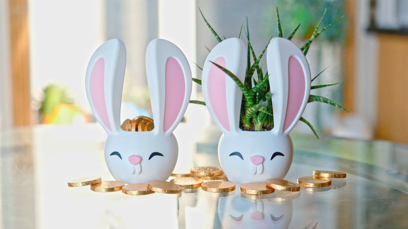 May include: Two white bunny-shaped planters with pink ears. One planter contains a small green plant, and the other has gold coins. Both planters are on a glass table with more gold coins scattered around them.