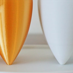 Puede incluir: Primer plano de dos macetas modernas y minimalistas. Una es de color dorado met&aacute;lico, la otra es blanca. Ambas tienen una base &uacute;nica de tres patas. Las macetas son probablemente para uso en interiores o exteriores.