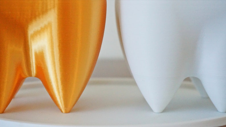 Puede incluir: Primer plano de dos macetas modernas y minimalistas. Una es de color dorado met&aacute;lico, la otra es blanca. Ambas tienen una base &uacute;nica de tres patas. Las macetas son probablemente para uso en interiores o exteriores.