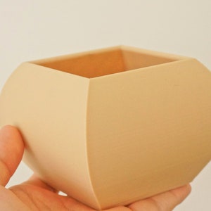 May include: A beige, square-shaped planter with rounded sides. The planter is held in a hand and has a smooth, matte finish. The interior is visible, revealing a lighter shade of beige. This decorative item is suitable for small plants.