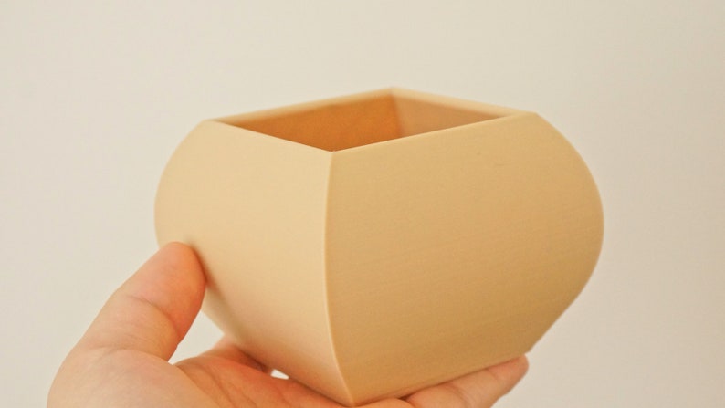 May include: A beige, square-shaped planter with rounded sides. The planter is held in a hand and has a smooth, matte finish. The interior is visible, revealing a lighter shade of beige. This decorative item is suitable for small plants.