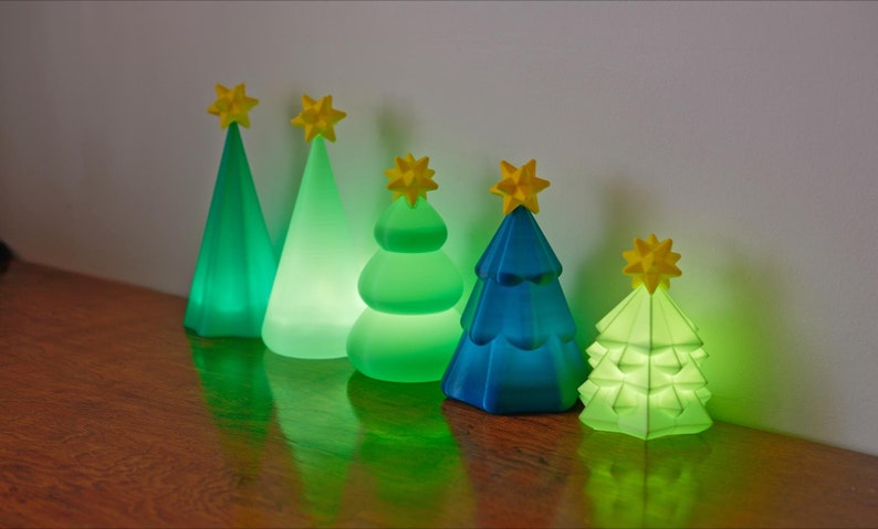 May include: Five illuminated Christmas tree decorations in varying shades of green and blue, each topped with a yellow star. The trees have different designs, including cone and layered shapes. The lights are on, creating a warm glow.