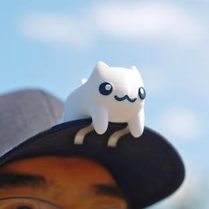 May include: A white cat-shaped accessory with black eyes and a smiling mouth sits on the brim of a dark blue baseball cap. The background is a bright blue sky. The accessory is a whimsical, decorative item.