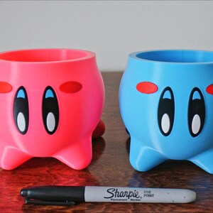 May include: Two 3D printed planters shaped like the character Kirby from the video game series Kirby. The planter on the left is pink and the planter on the right is blue. Both planters have black eyes, white pupils, and red blush.
