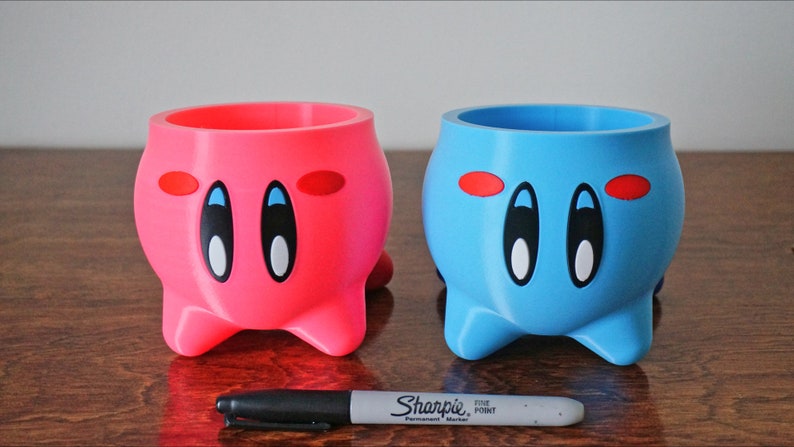 May include: Two 3D printed planters shaped like the character Kirby from the video game series Kirby. The planter on the left is pink and the planter on the right is blue. Both planters have black eyes, white pupils, and red blush.