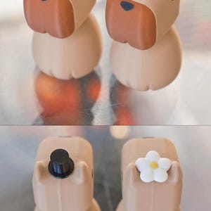 May include: Two small, beige, 3D printed figurines of dogs. The dog on the left is wearing a black top hat. The dog on the right is wearing a white flower on its head.
