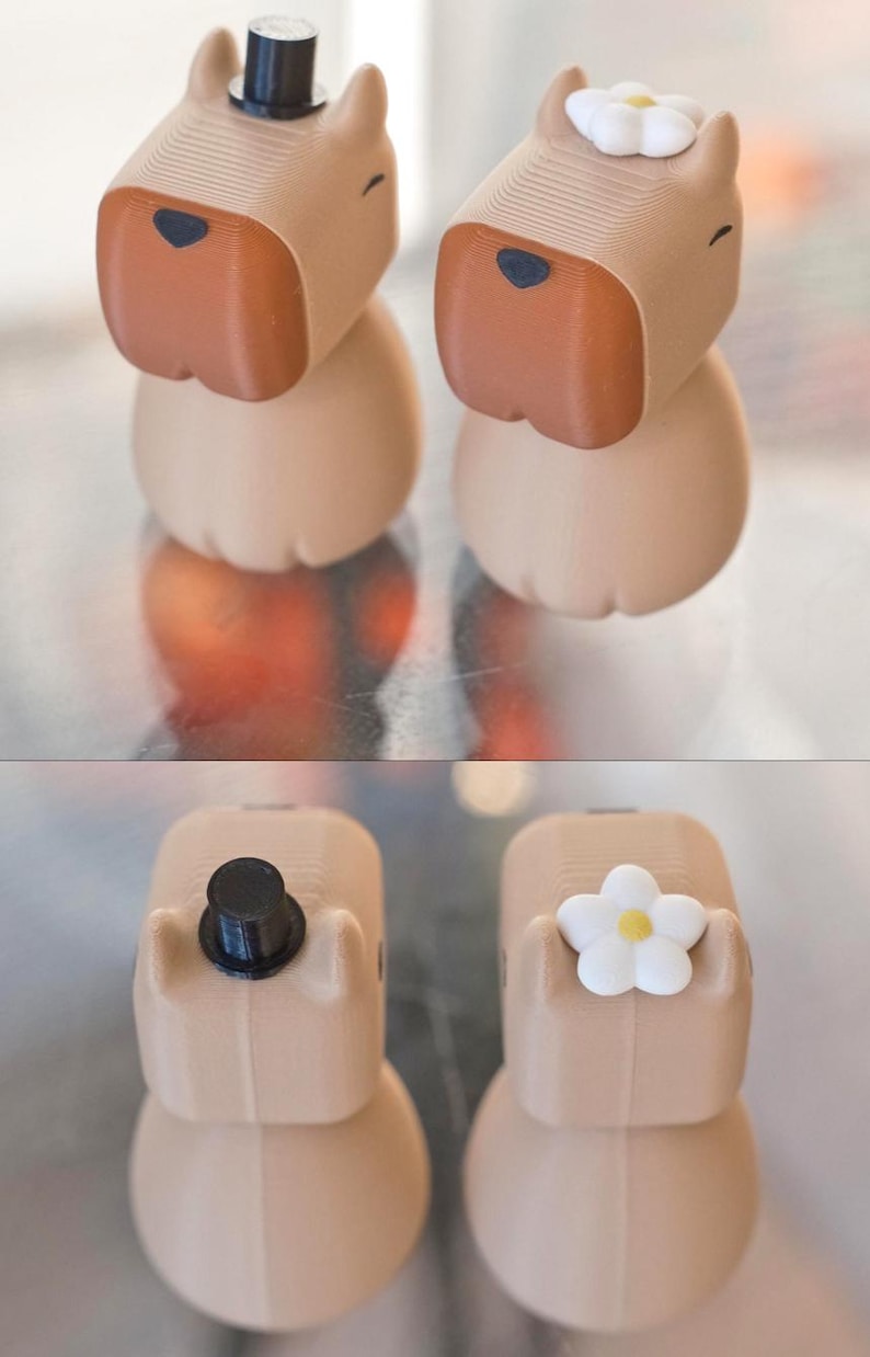 May include: Two small, beige, 3D printed figurines of dogs. The dog on the left is wearing a black top hat. The dog on the right is wearing a white flower on its head.