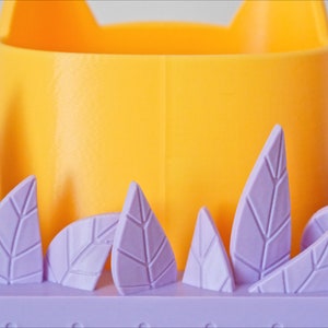 Peut inclure: Un pot de fleurs en forme de chat jaune avec une base violette et des feuilles violettes autour du bord.