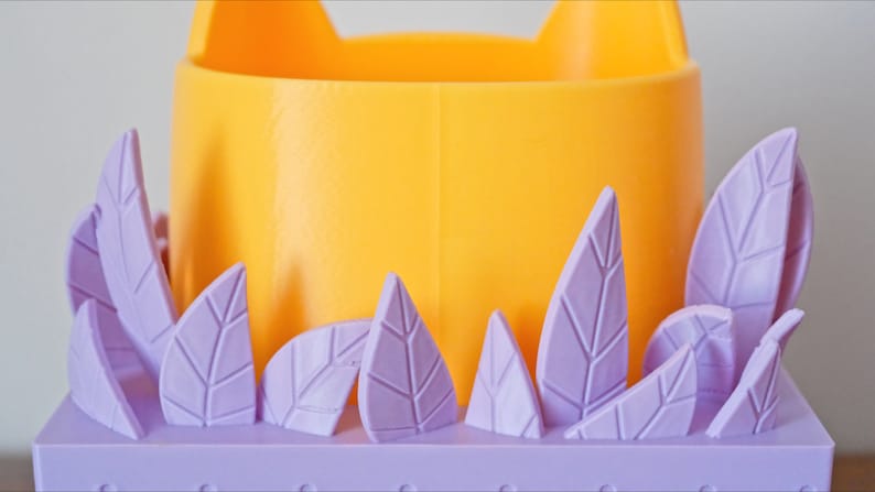 Peut inclure: Un pot de fleurs en forme de chat jaune avec une base violette et des feuilles violettes autour du bord.